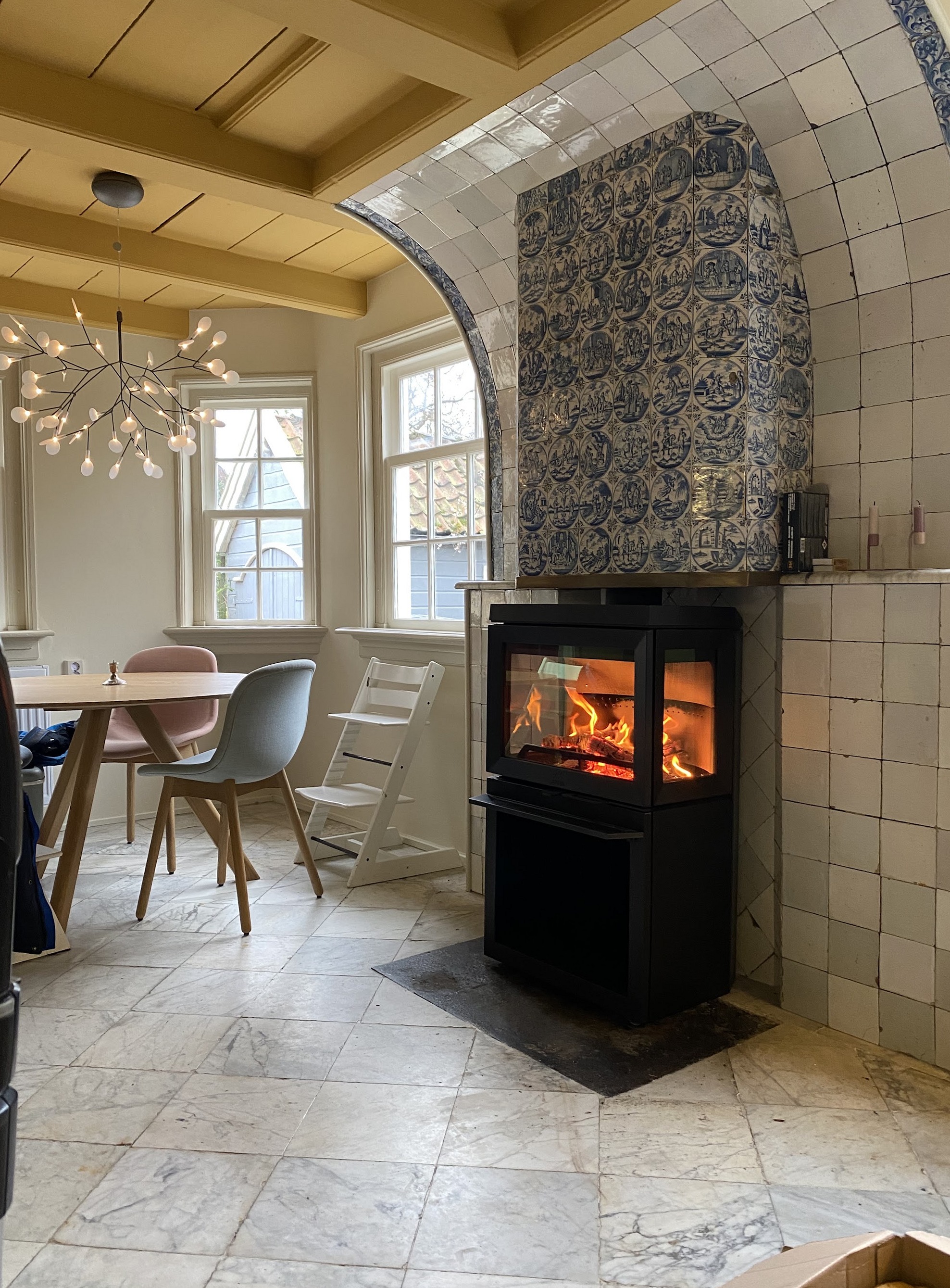 Een brandende houtkachel in een nis met Delfts blauwe tegels, in een keuken met een eettafel.