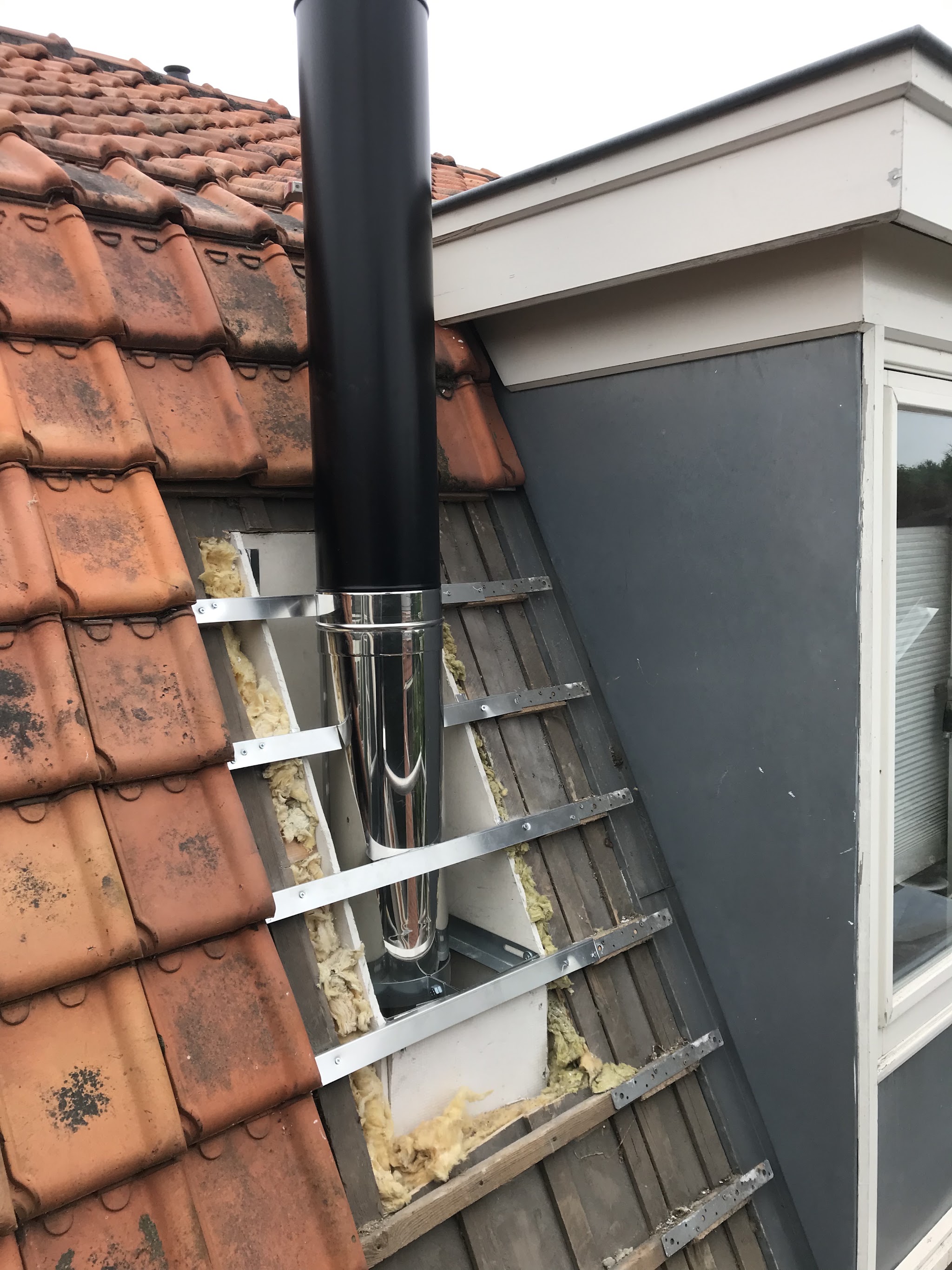 Een zwarte rookkanaalpijp die door een rood pannendak naar buiten komt, naast een dakkapel.