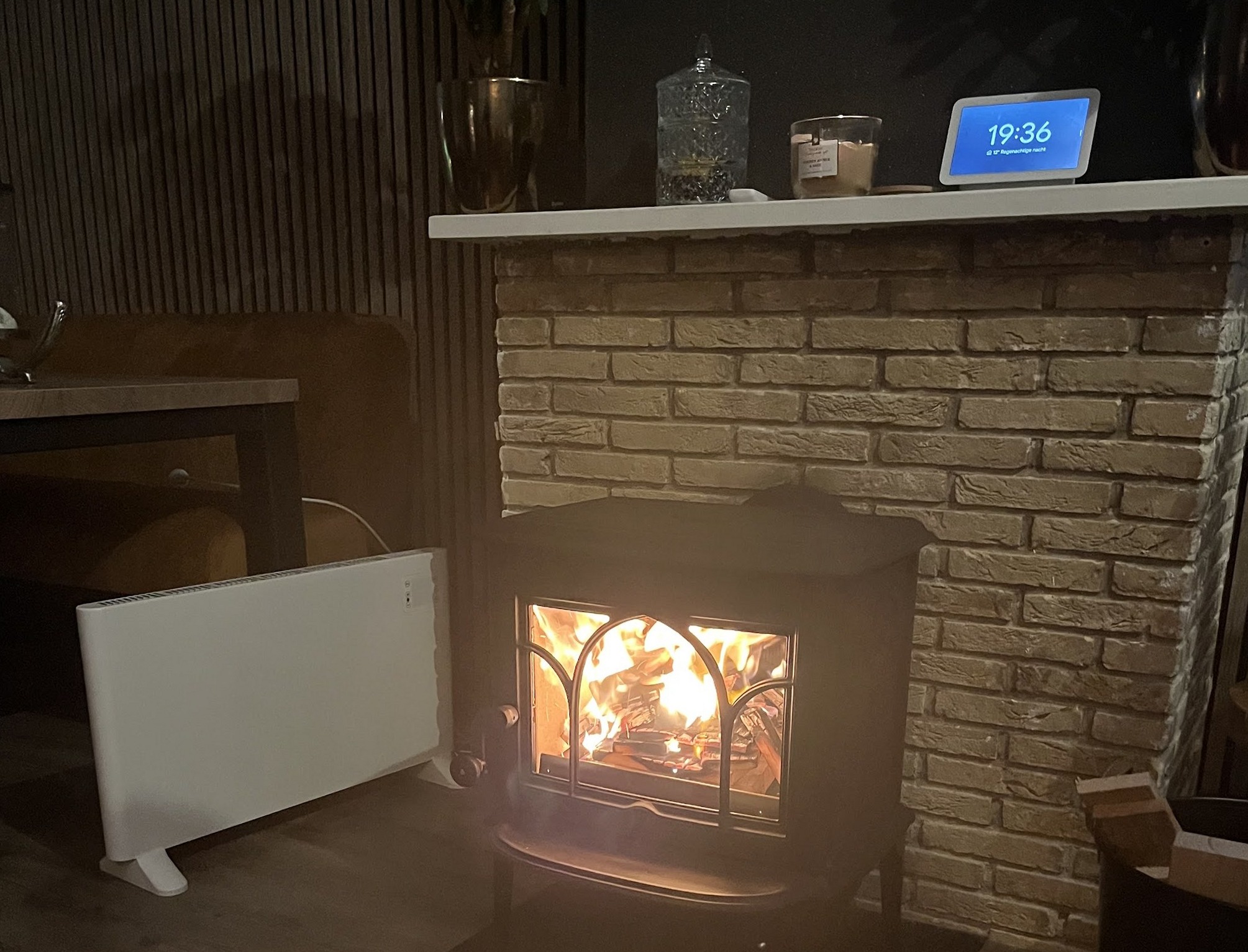Een brandende houtkachel voor een lichte bakstenen schoorsteenwand met een schouw erboven.